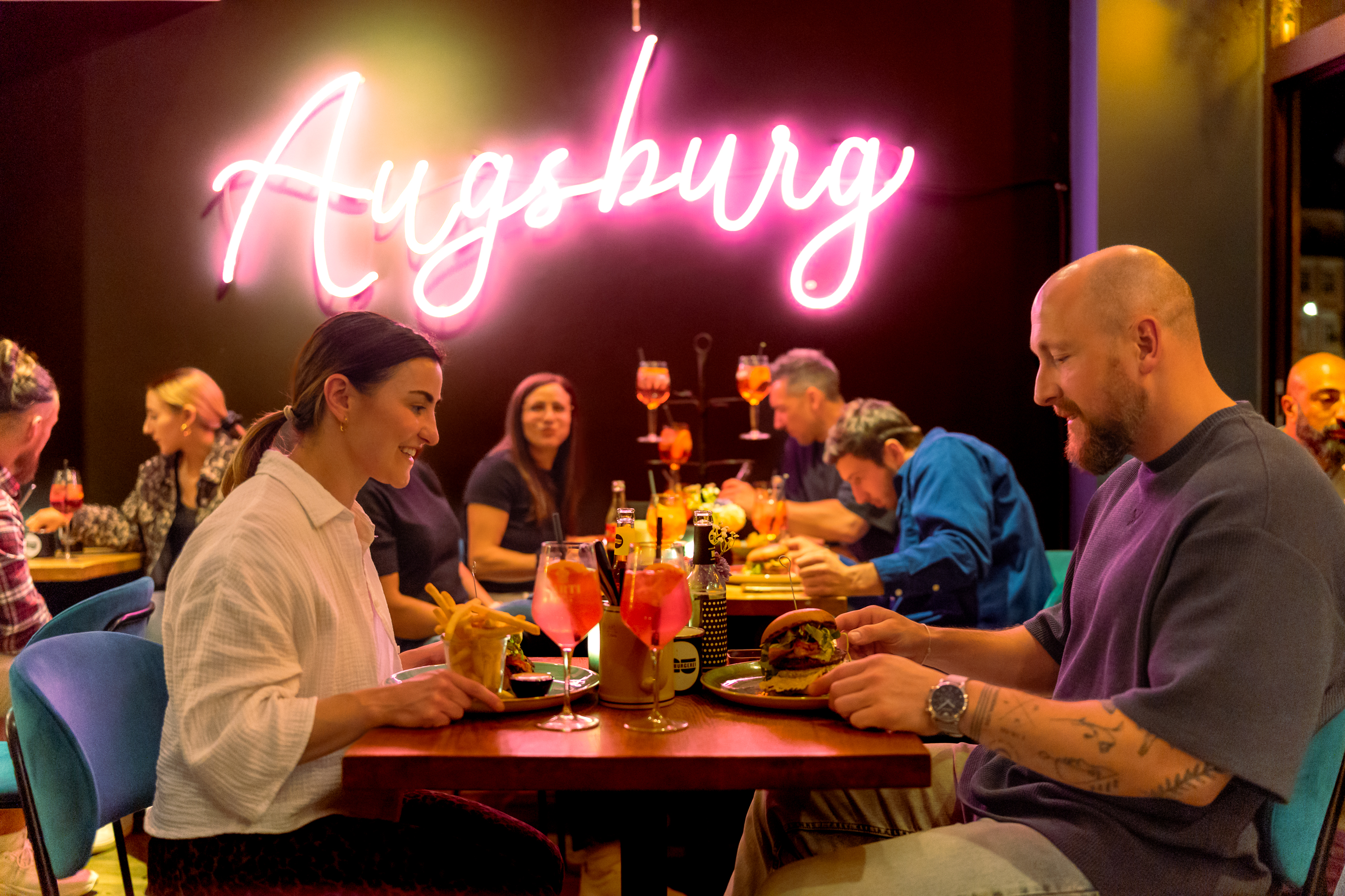 Gäste genießen Burger unter dem Augsburg-Neonschriftzug in der Hamburgerei